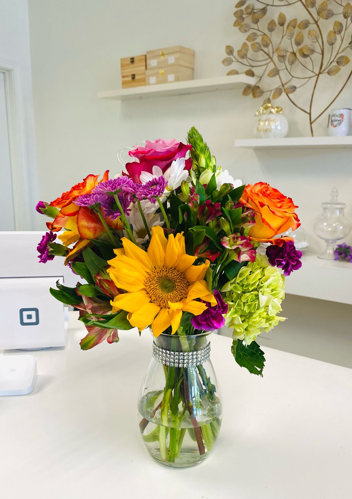 Vibrant bouquet of sunflowers and roses in a glass vase on a white desk with shelves in background.