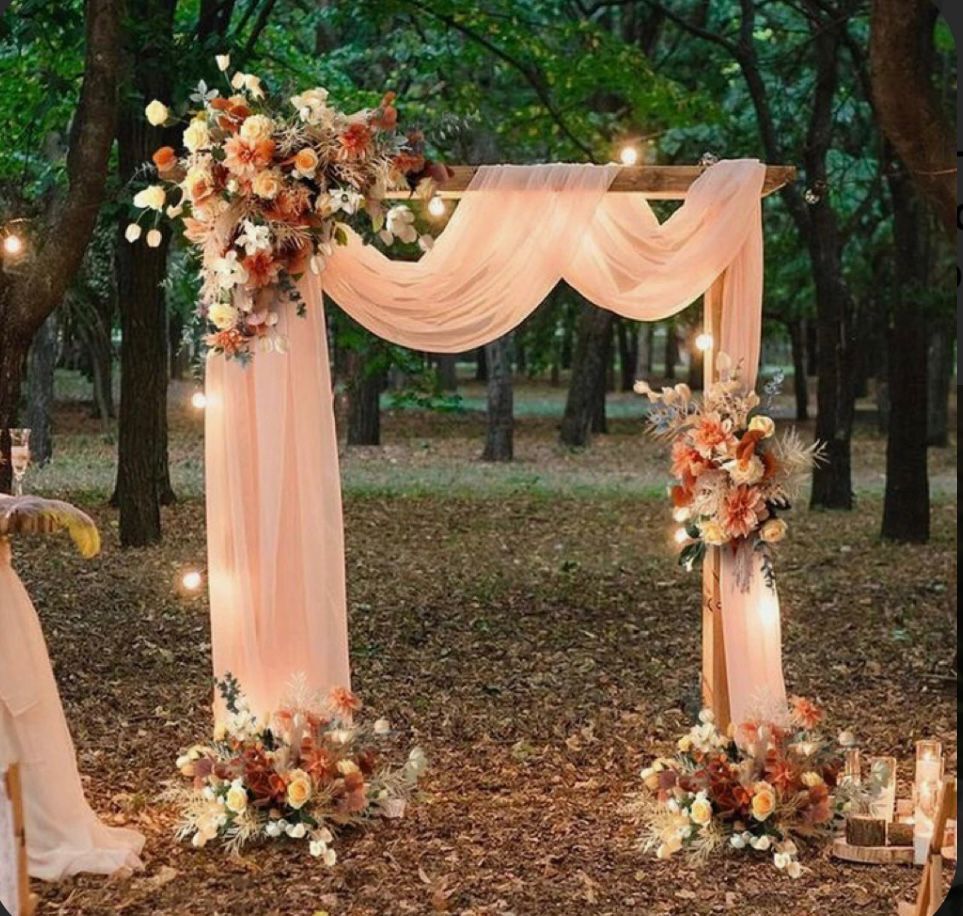 Wedding arch decorated with flowers and sheer peach fabric, set in a wooded area with string lights.