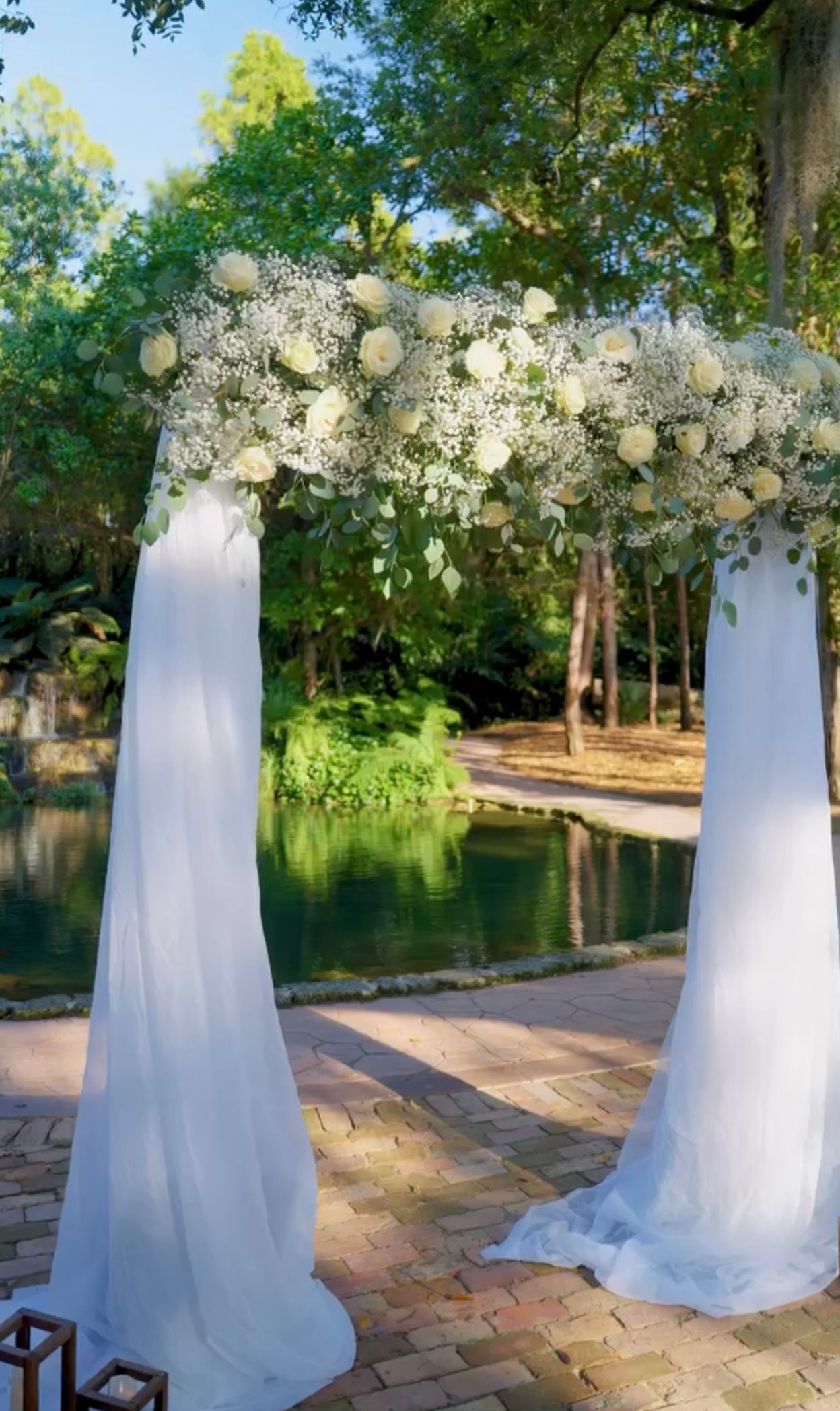 Wedding arch draped with white fabric and flowers, in front of a pond and trees.
