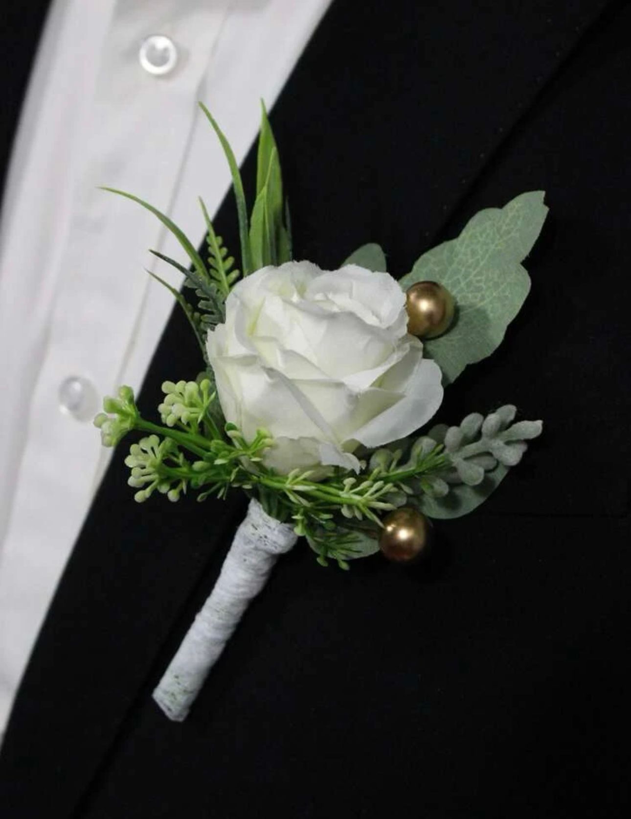 White rose boutonniere pinned to a black suit, with green accents and gold beads.