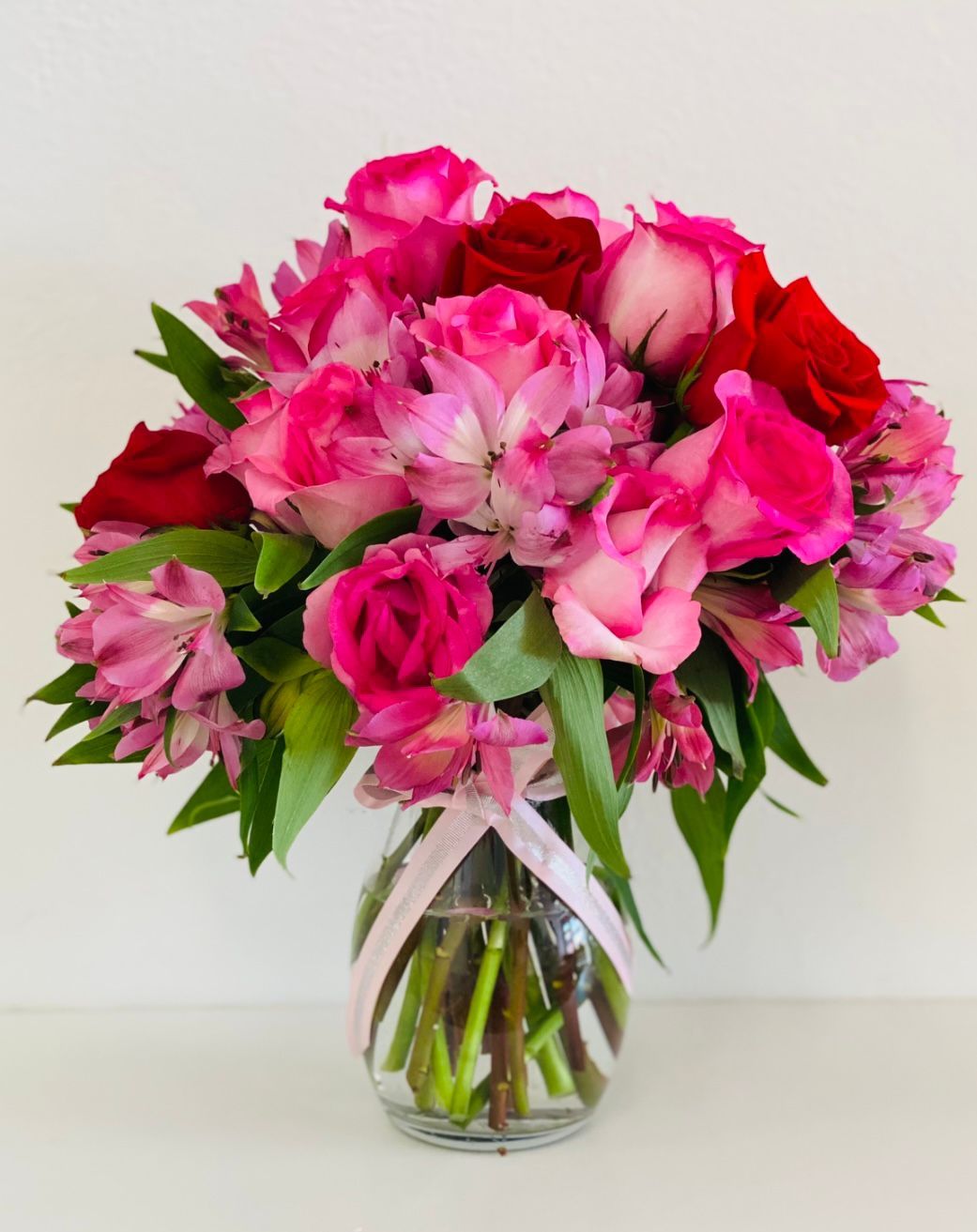 A bouquet of pink and red roses and alstroemeria in a clear glass vase with a pink ribbon.
