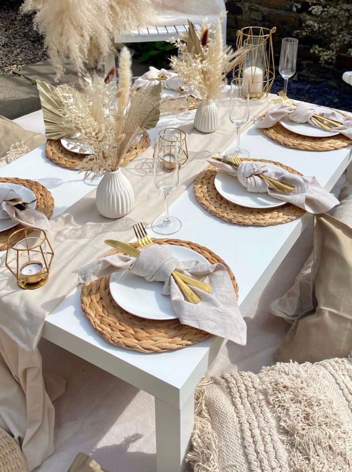 Picnic table with neutral decor: white, beige, gold. Pampas grass, woven placemats, folded napkins, candles. Outdoor setting.
