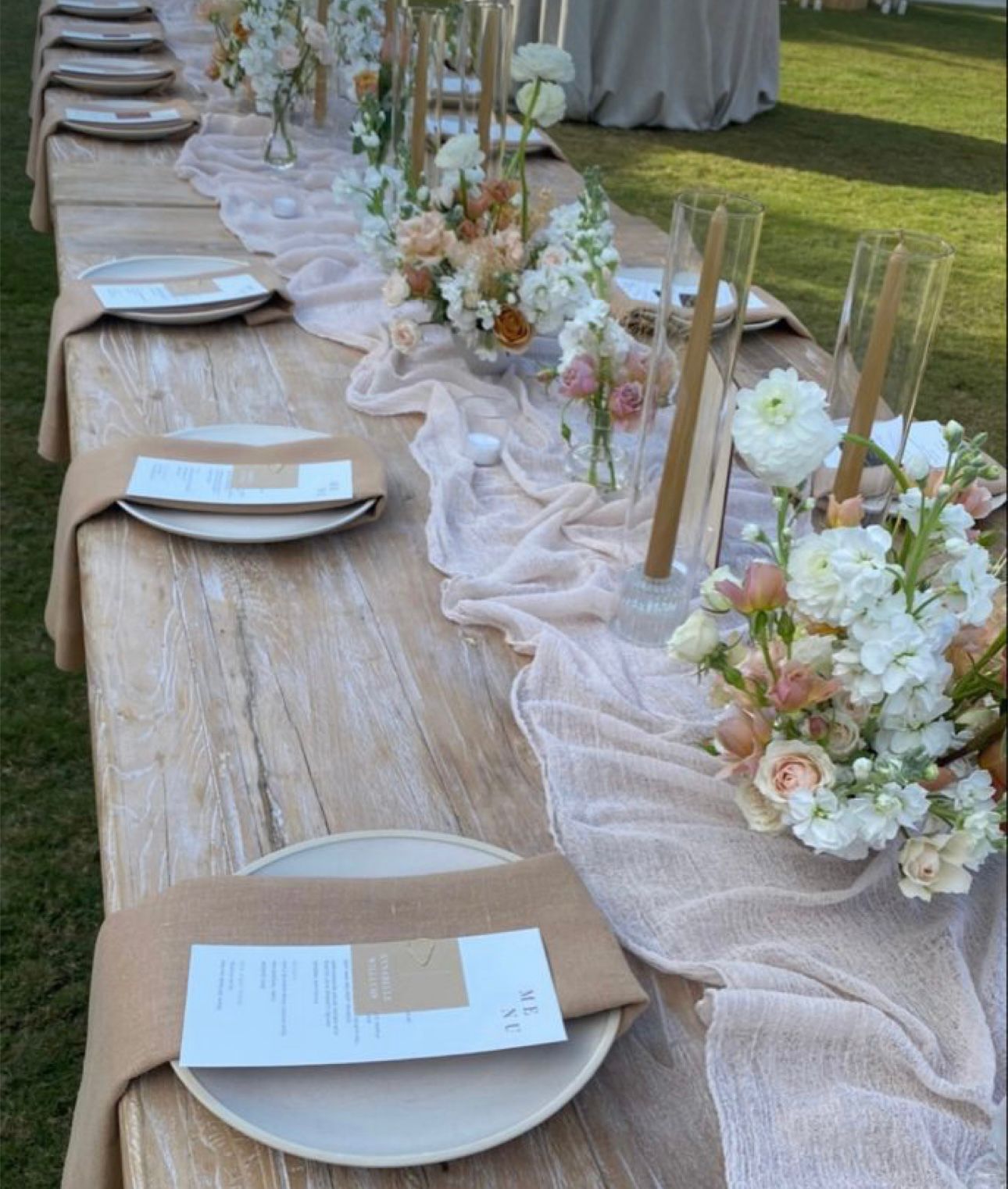 Long wooden table set for an outdoor event; tan tablecloth with flower arrangements, plates, and menus.