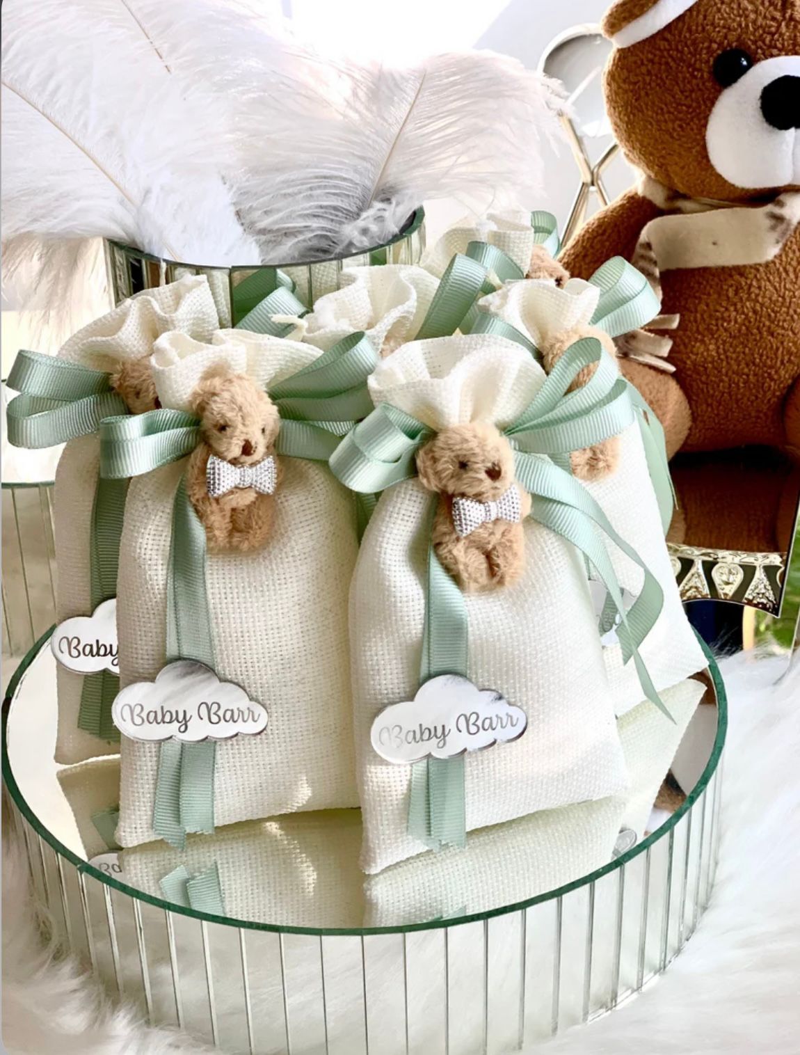 Close-up of baby shower favors. White bags with green ribbon and teddy bear charms, set on a mirrored tray.