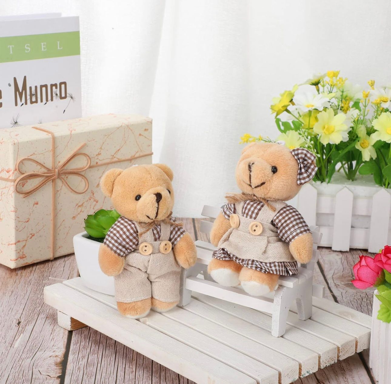 Two tan teddy bears with plaid outfits sit on a small white bench; flowers and gift box in background.