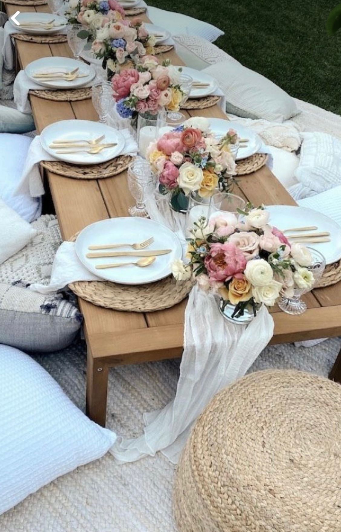 Picnic setup: low wooden table with flowers, place settings, pillows, and woven mats on the grass.