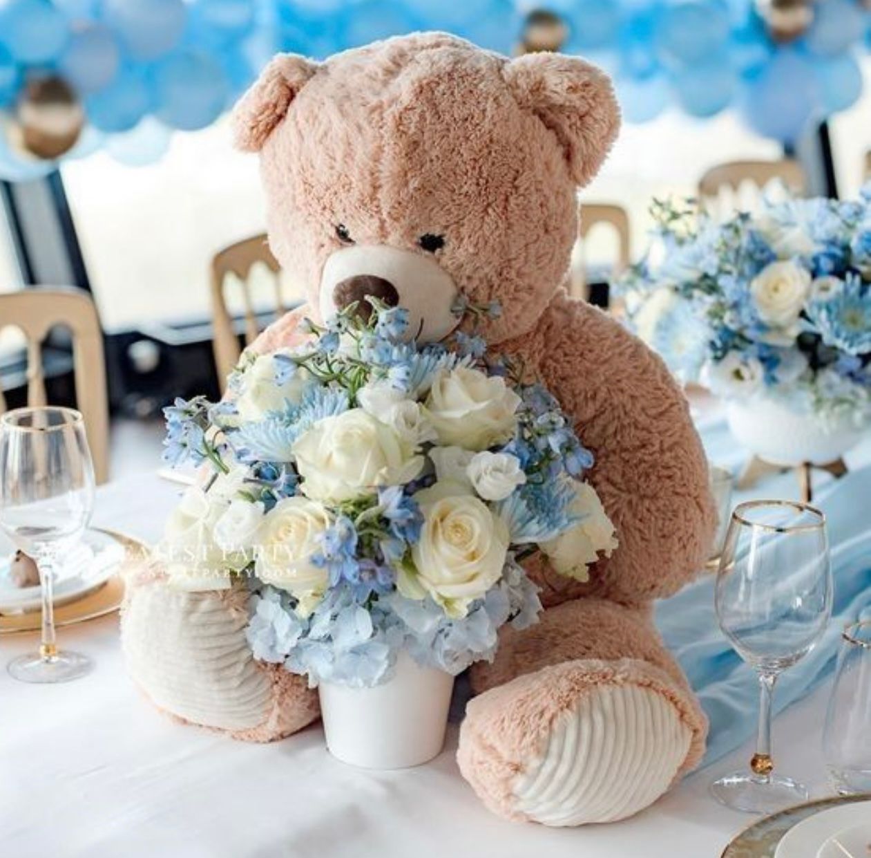 Large teddy bear holding a bouquet of white and blue flowers, set on a table decorated for an event.