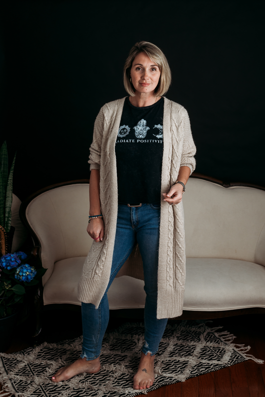 Shannon A Fisher  standing barefoot in a cozy room, wearing a black T-shirt, jeans, and a cream cardigan in front of a sofa