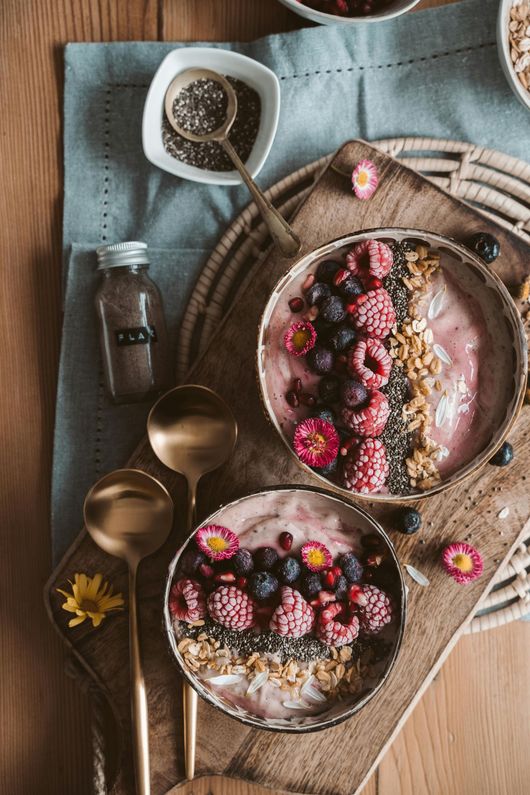 Two berry smoothie bowls with chia seeds, nuts, and flowers on a wooden table with spoons and a bottle
