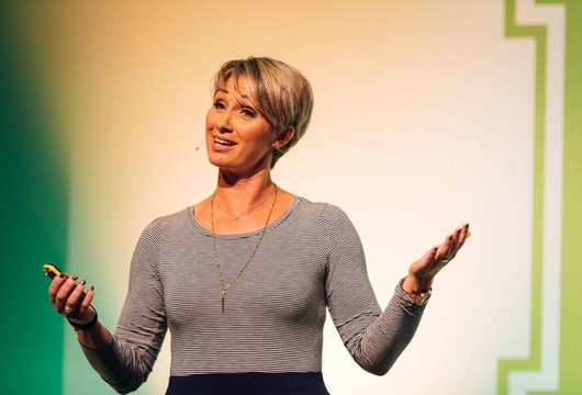 Shannon A Fisher  speaking onstage in a striped shirt, gesturing with both hands against a green and white backdrop