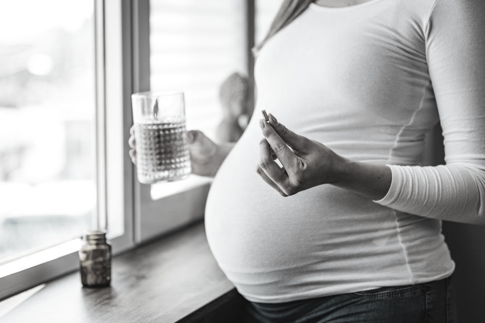 Pregnant person holds a pill and glass of water by a window, near a bottle.