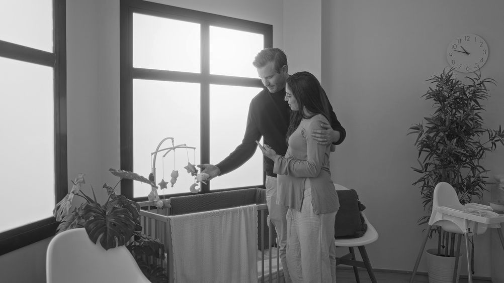 Couple in nursery, man pointing at mobile above crib, woman holds phone, sunny room.