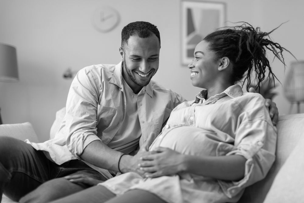 Smiling couple, pregnant woman holding belly, man's arm around her on a couch.