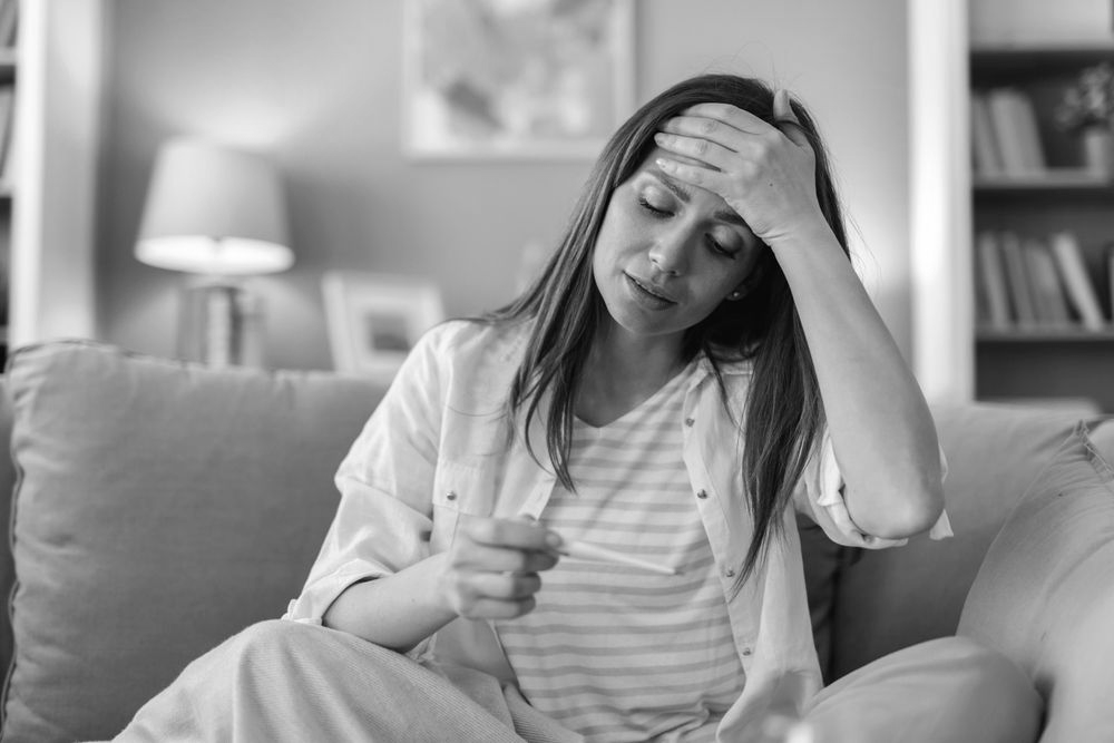 Woman on couch holding thermometer and forehead in hand, looking unwell in a living room
