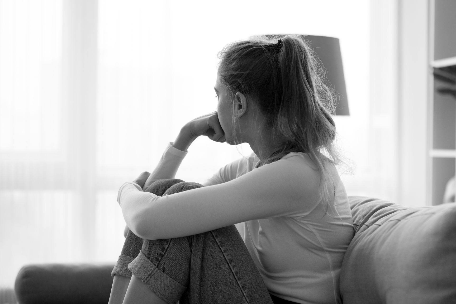 A nervous young woman sitting on a couch and staring out a window.