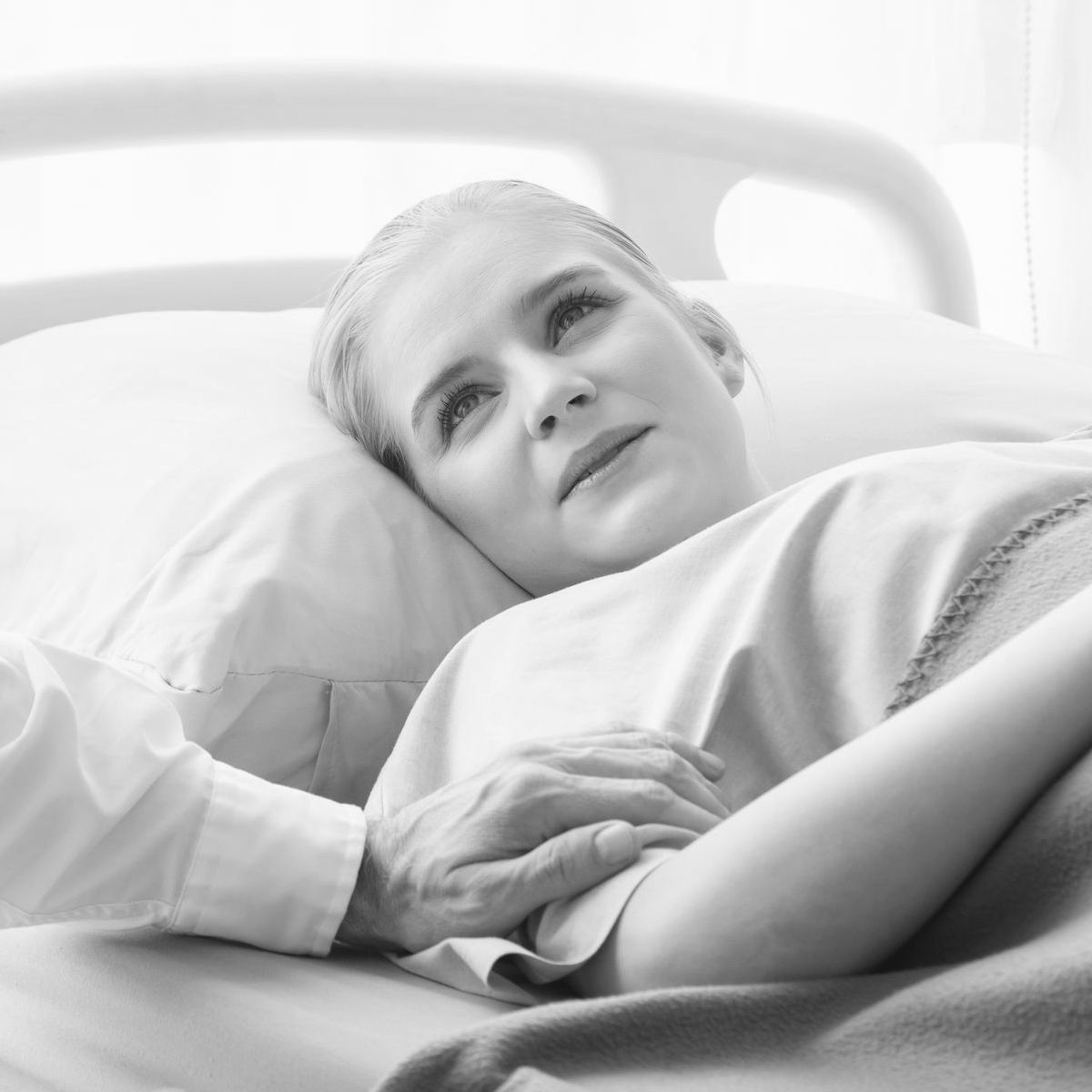 Woman lying in a hospital bed looking up while someone gently places a comforting hand on her arm.