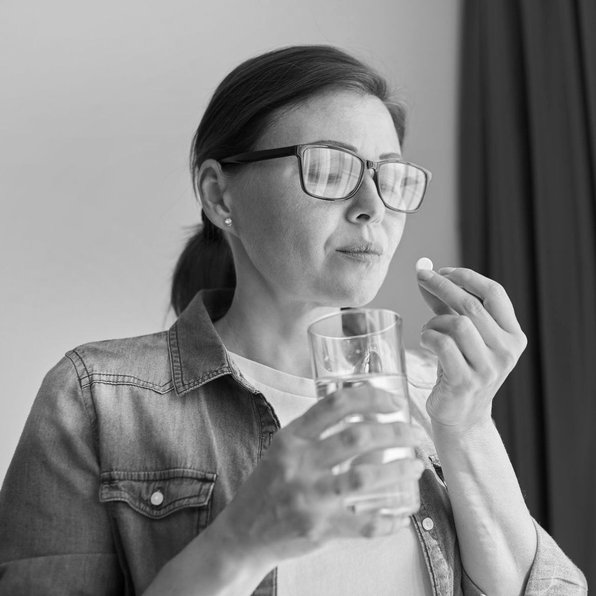 Woman wearing glasses holding a glass of water and preparing to take a pill.