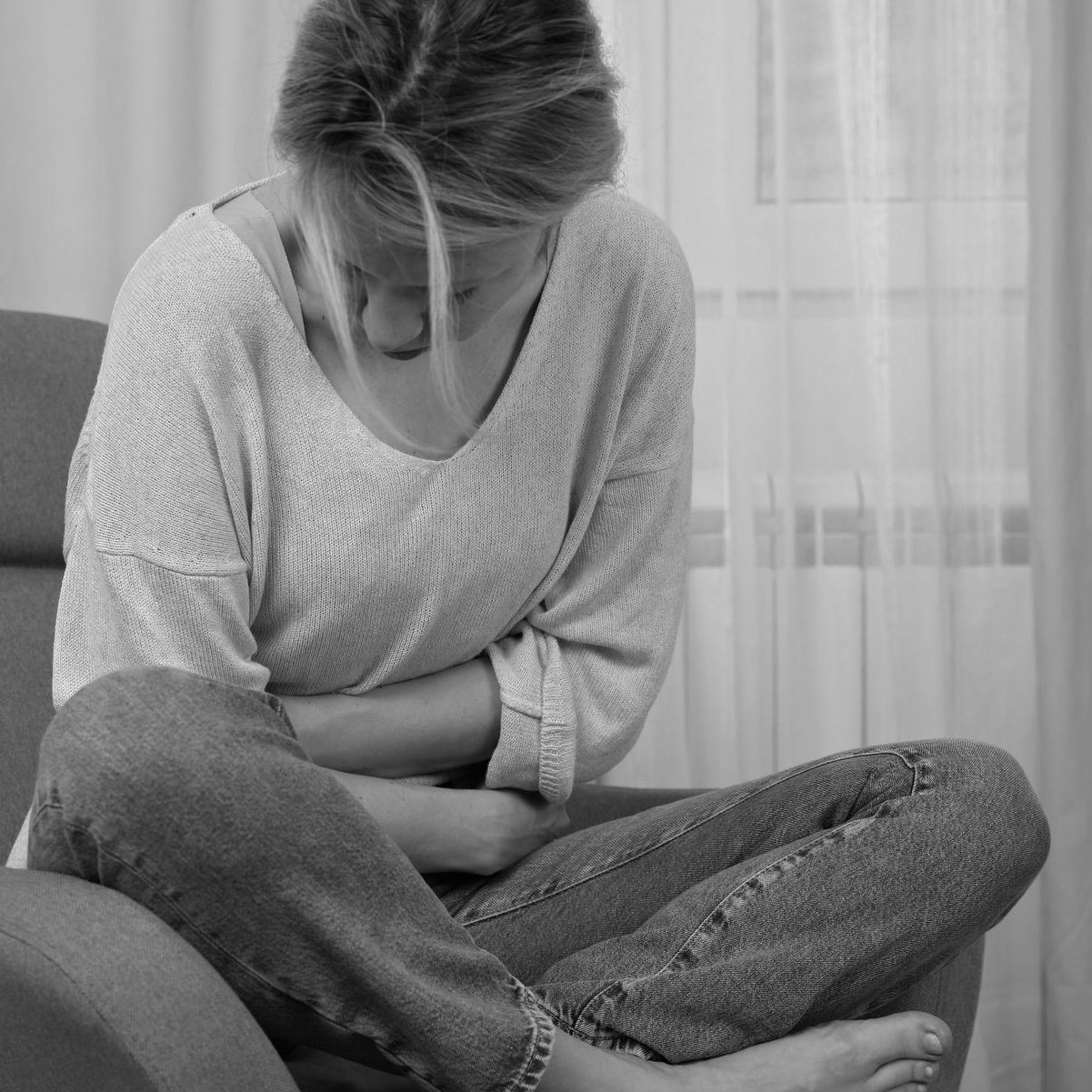 Woman sitting on a chair holding her stomach in discomfort, appearing to have abdominal pain.