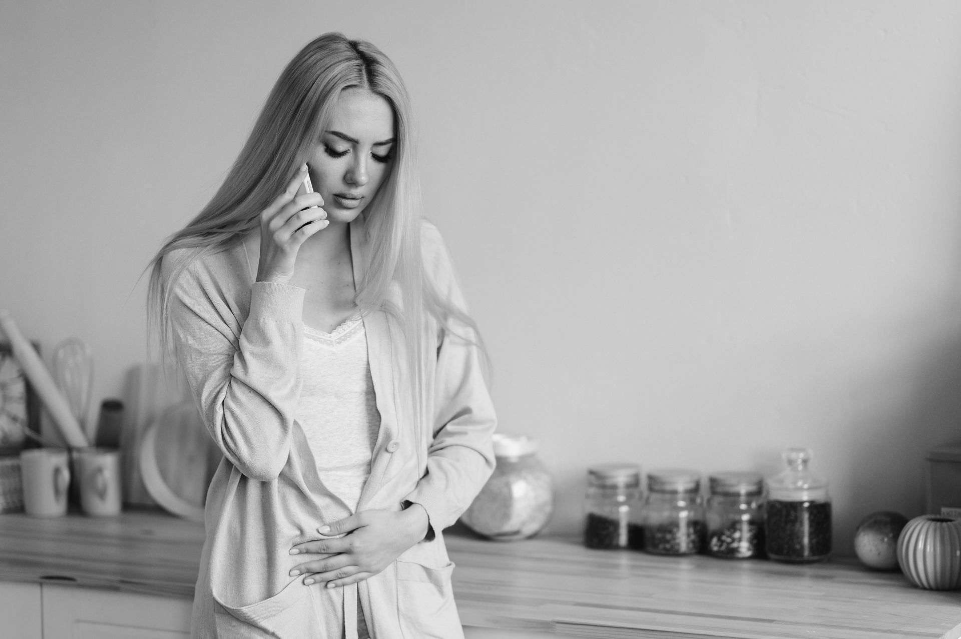 Worried woman standing in a kitchen, holding her stomach while talking on the phone.