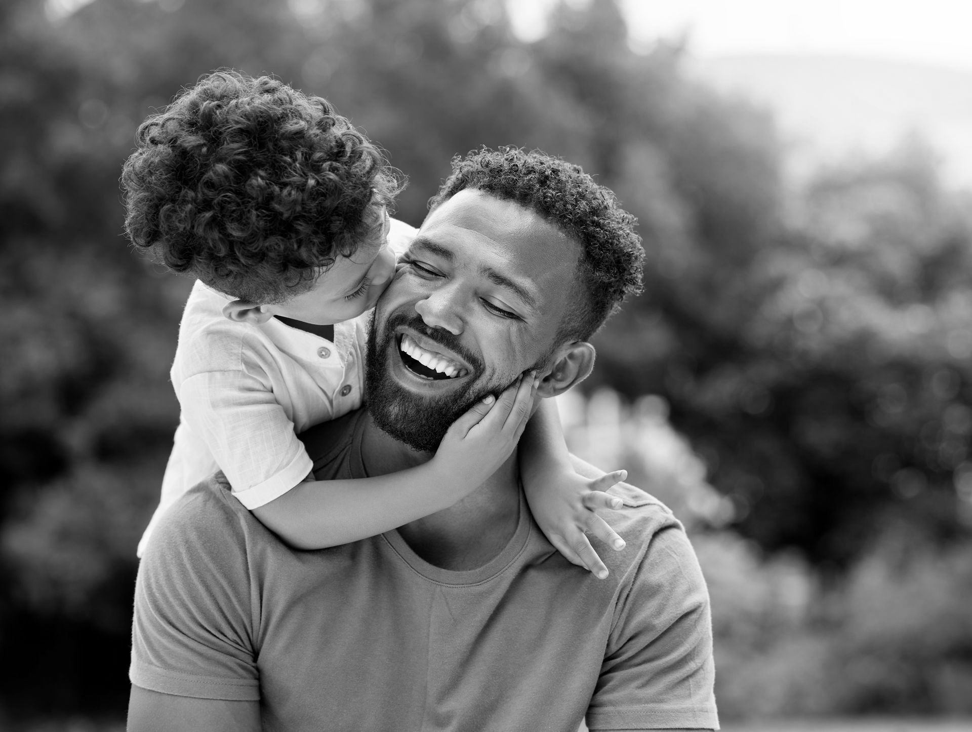 Laughing father holding his young child who is kissing him on the cheek outdoors.