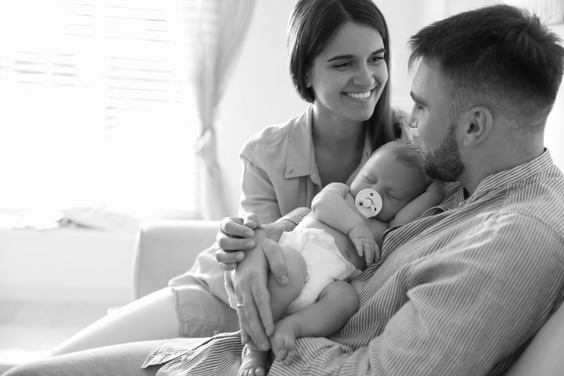 Smiling parents sitting together as the father holds their sleeping baby with a pacifier in his arms.