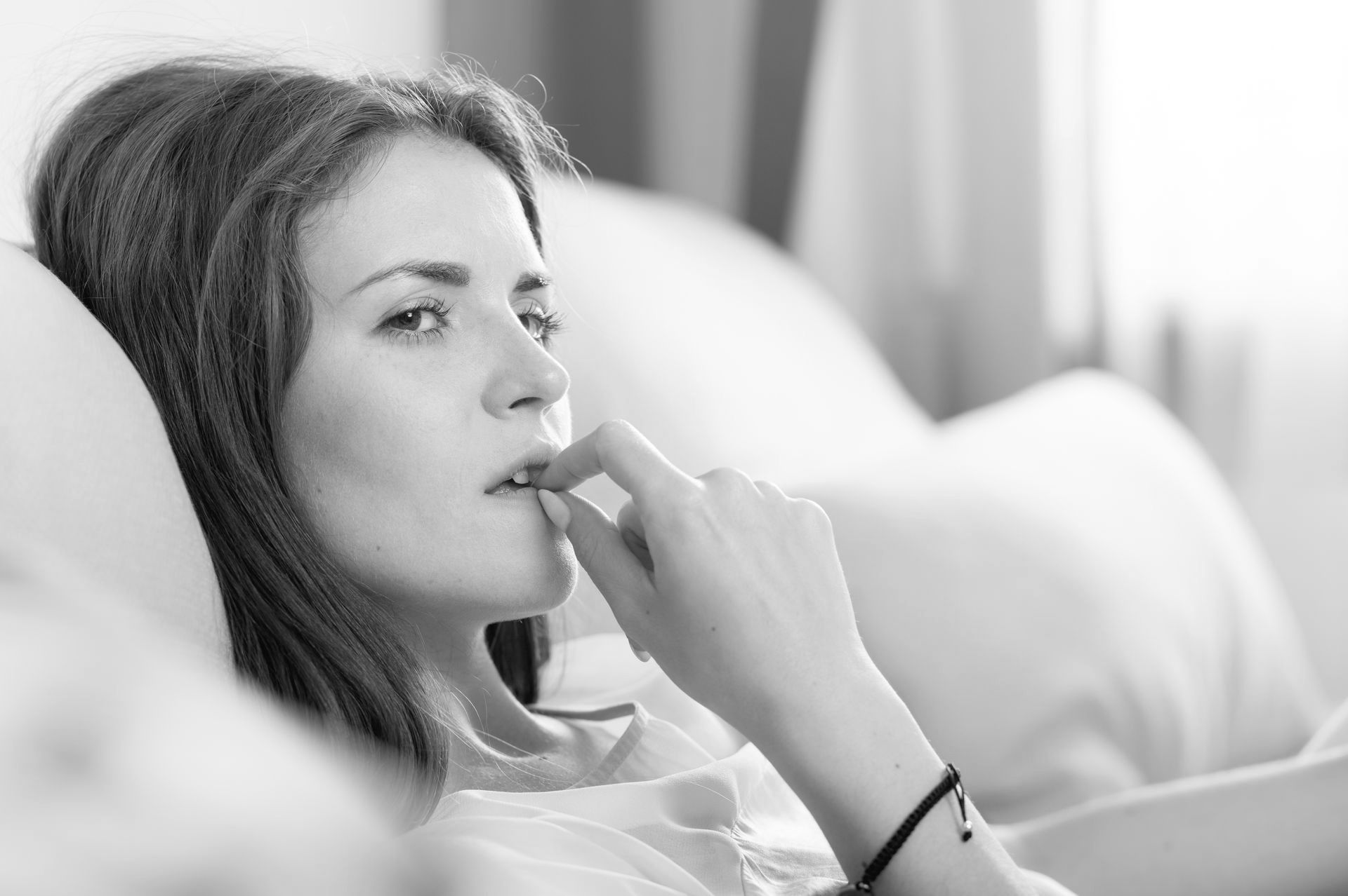Woman sitting on a couch, looking thoughtful and concerned while resting her hand near her mouth.