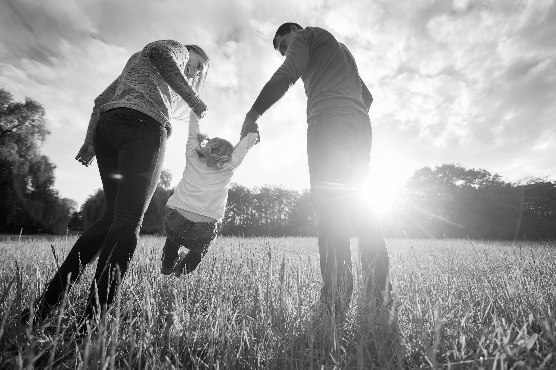 Parents swing their young child by the arms while walking through a sunny grassy field together.