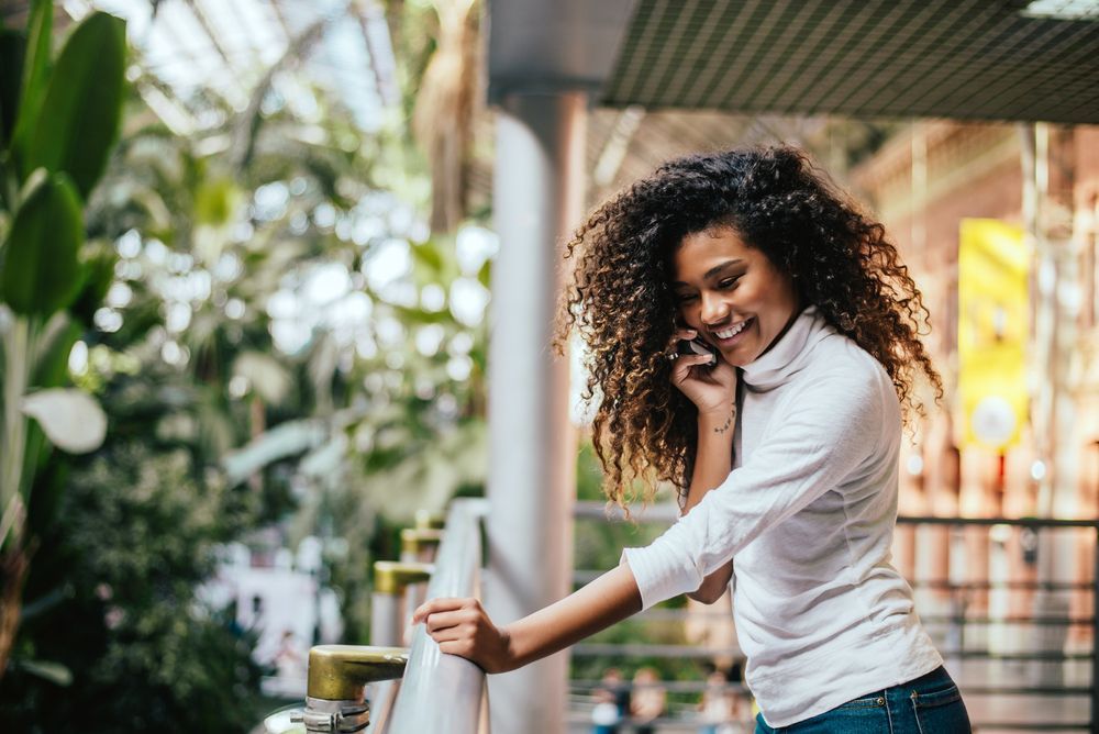Smiling woman talking on her phone while leaning on a railing in a bright, green indoor space.