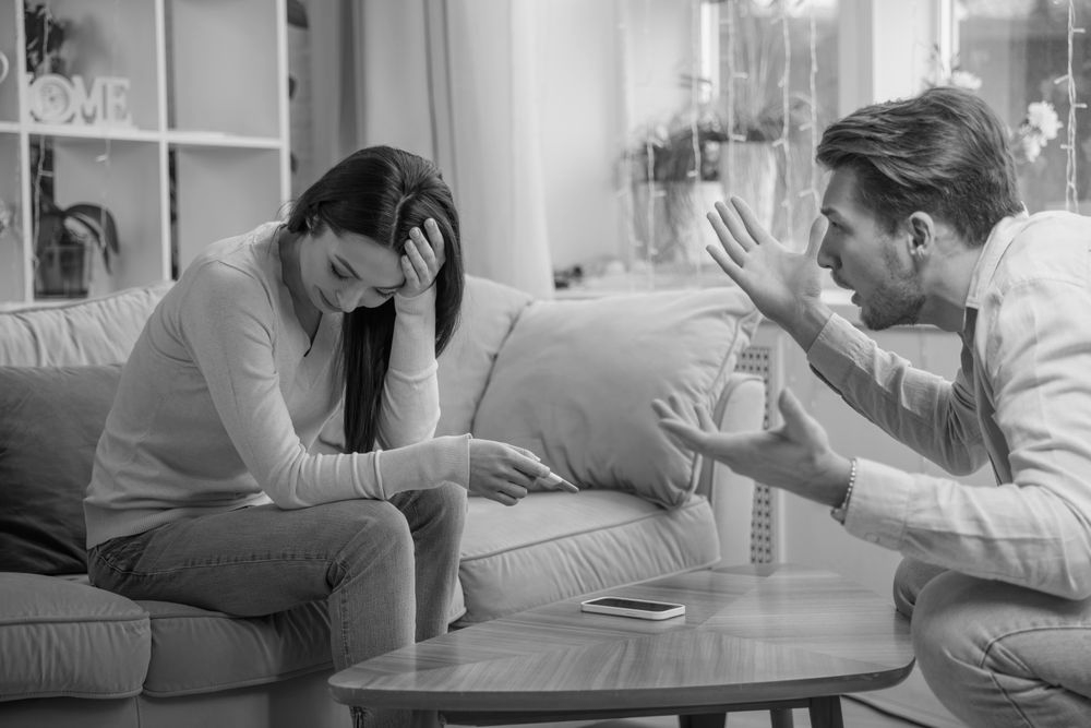 Two people arguing on a couch in a living room, one upset and looking down, the other gesturing emphatically.