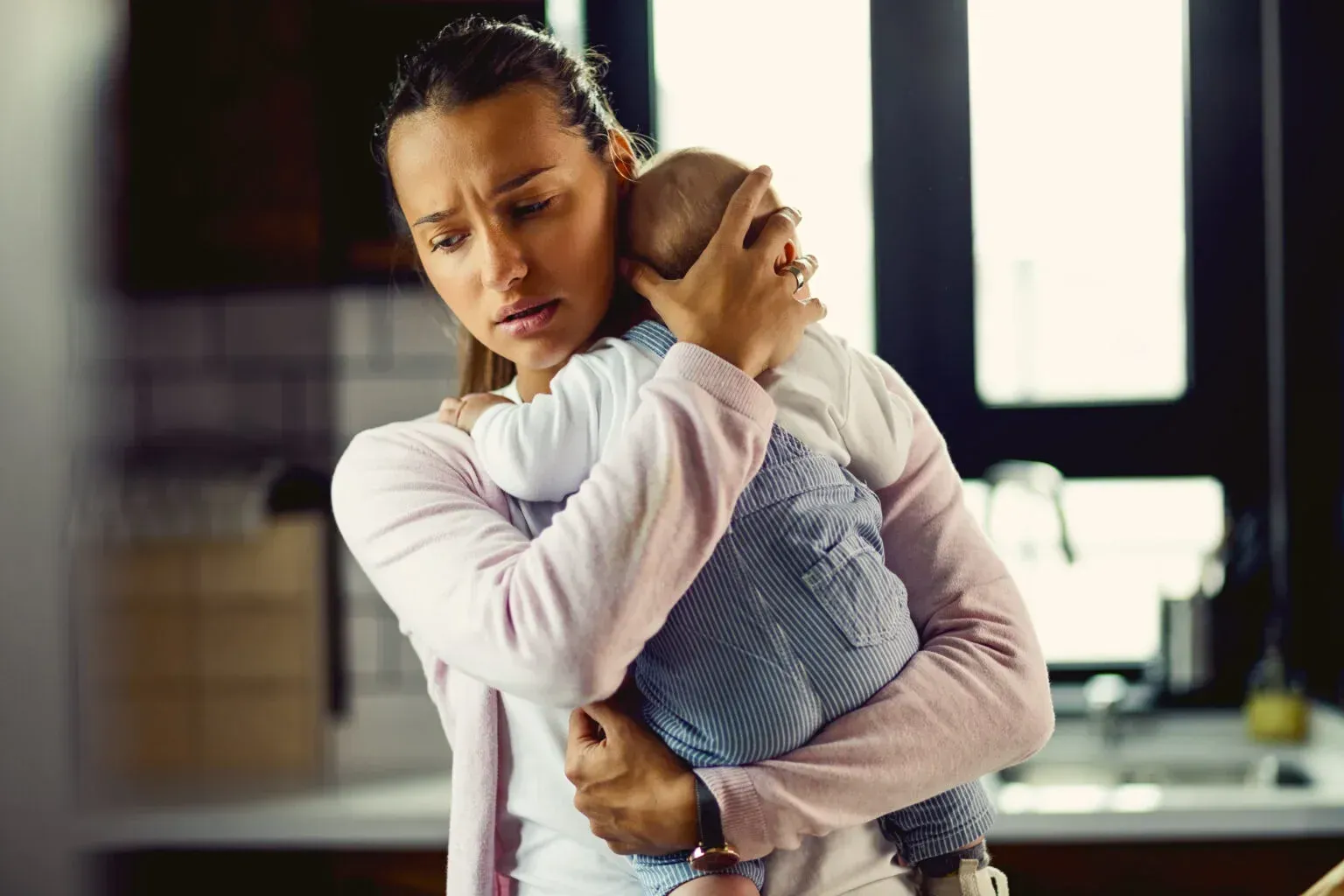 A woman suffering from post partum depression is holding a baby in her arms in a kitchen.