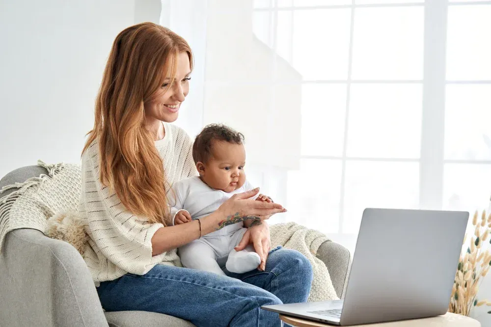 A woman is sitting on a couch holding a baby and using a laptop.