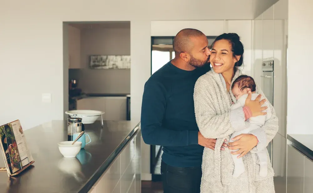 A man is kissing a woman on the cheek while holding a baby in a kitchen.