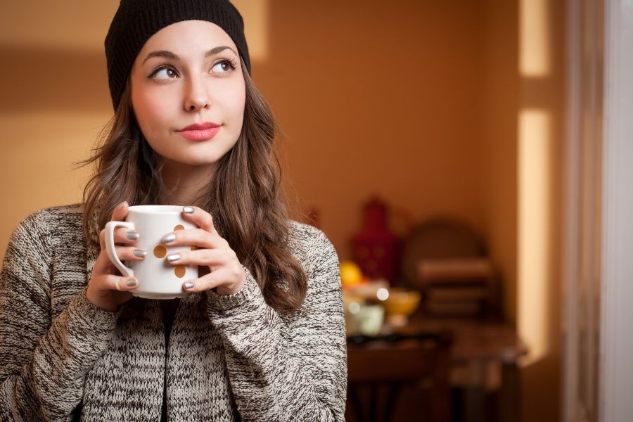 Thoughtful young woman wearing a beanie and sweater, holding a coffee mug and looking to the side.