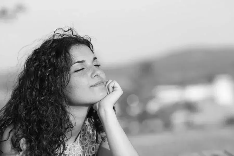 Woman with curly hair sitting outdoors with eyes closed, smiling peacefully in the sunlight.