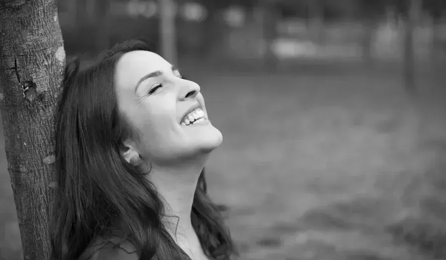 A woman is leaning against a tree in a park and smiling.