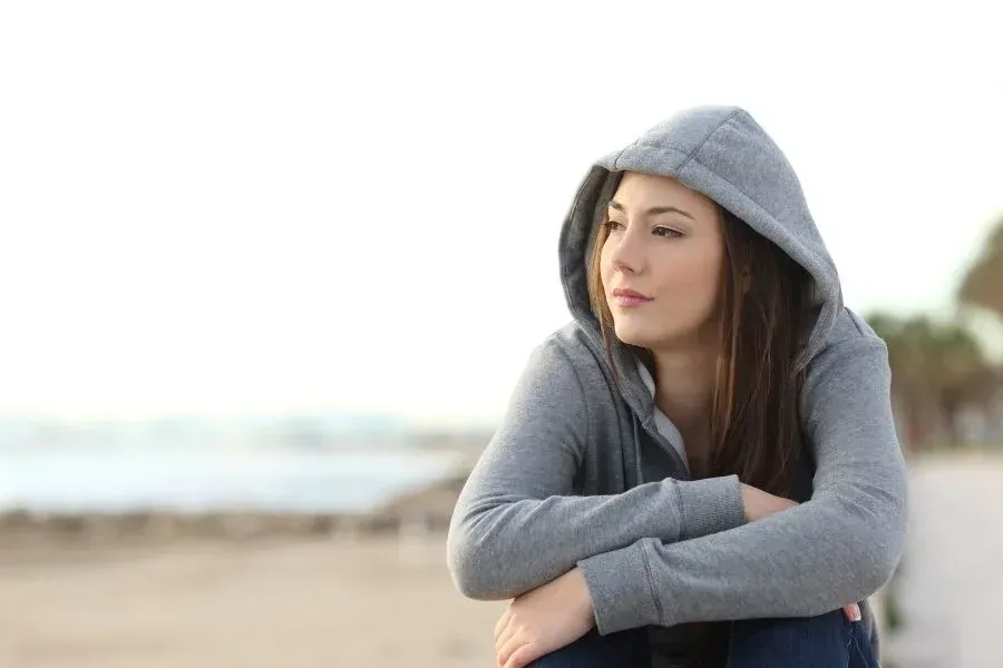 A woman in a hoodie is sitting on the beach looking at the ocean.