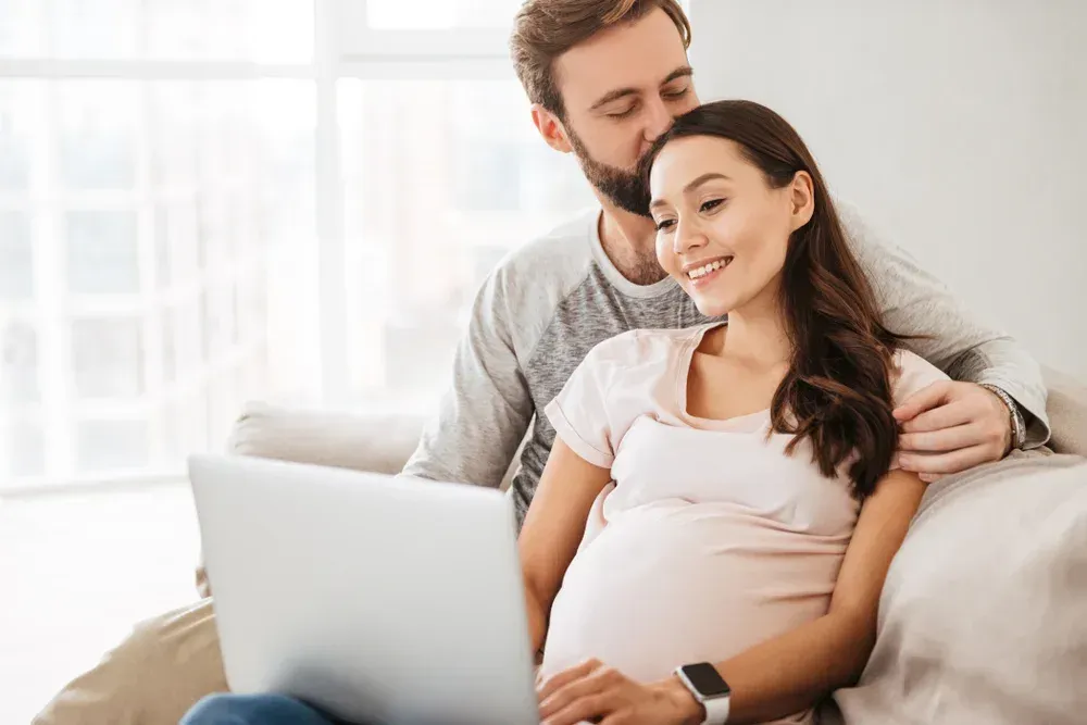 A young man relaxing with his pregnant wife on the couch while she uses a laptop.