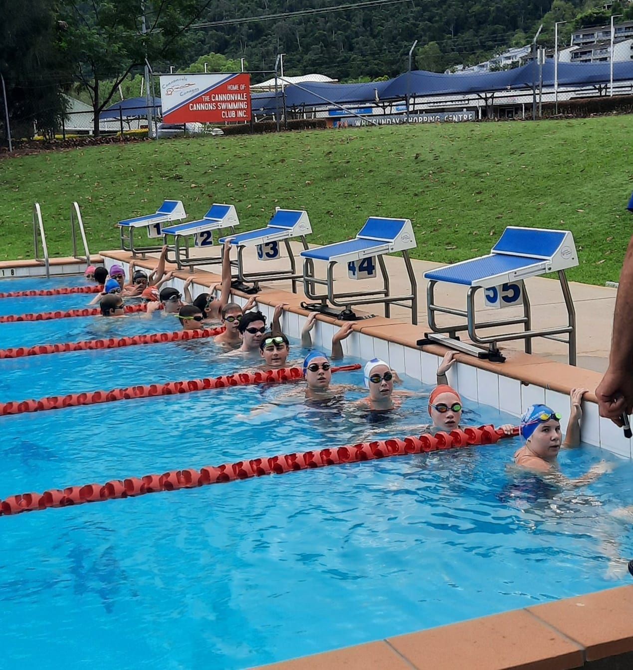Swimmers line up in pool lanes facing starting blocks at an outdoor competition.