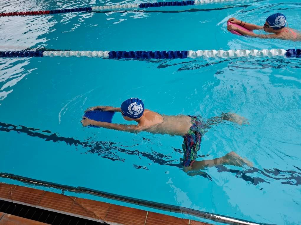 Two swimmers in blue caps practice with kickboards in separate lanes of an indoor pool.