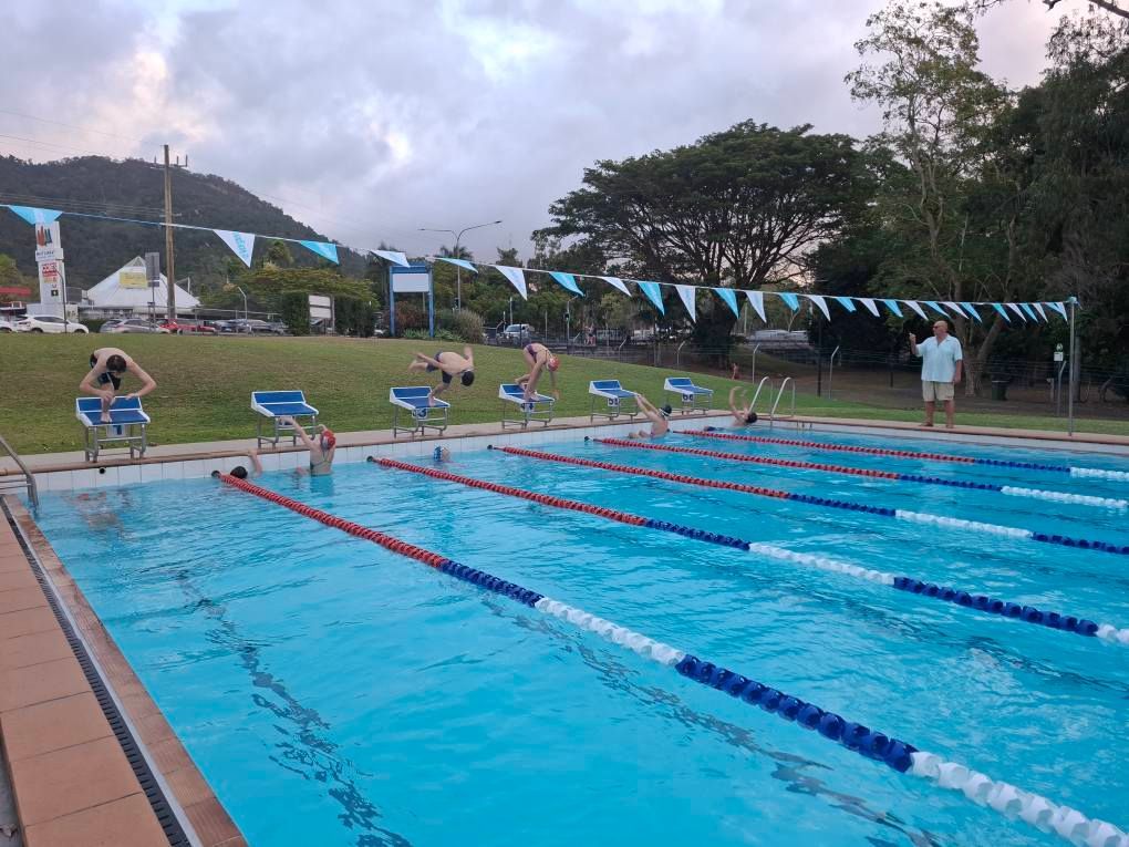 Swimmers at starting blocks, preparing to dive into a blue outdoor pool. Flags and mountains in the background.