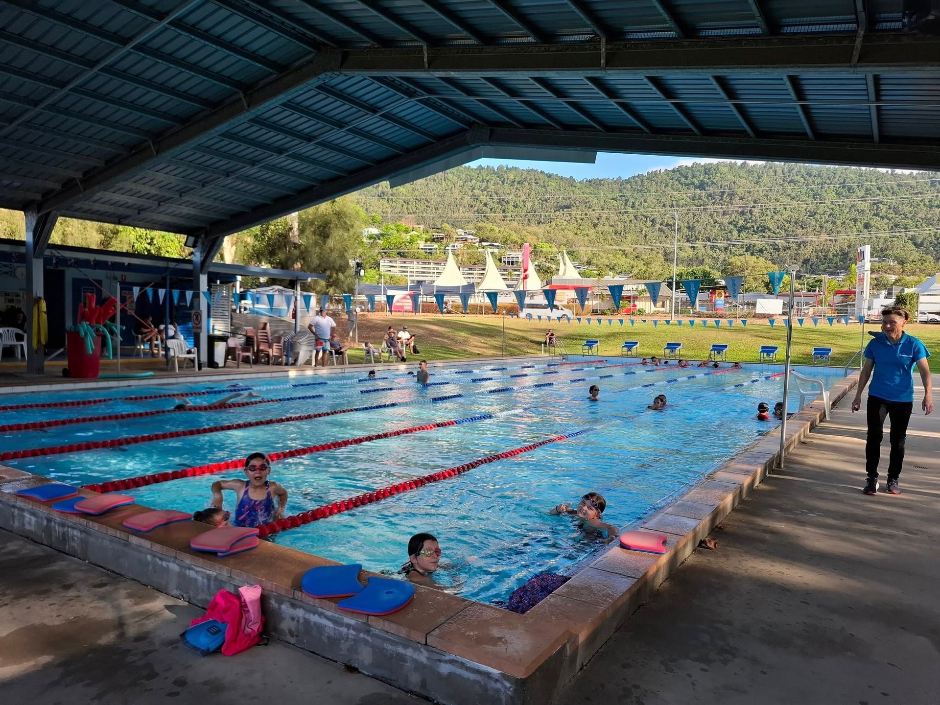 A covered outdoor pool with marked lanes, people swimming, and a person walking on the poolside.