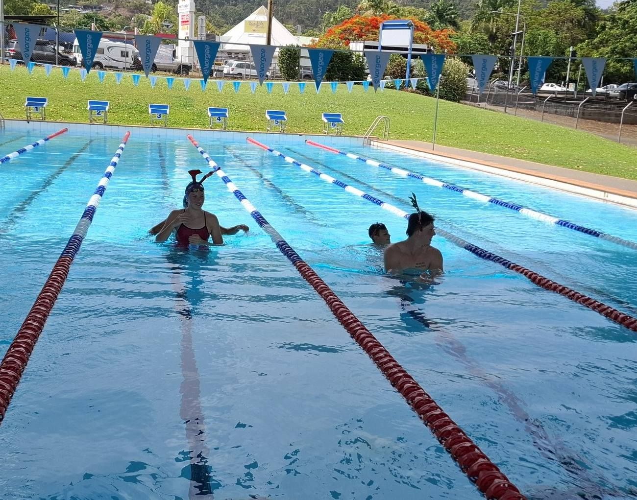 Swimmers in a Blue Outdoor Pool. Two Wear Headpieces, Others Swim — Cannonvale Swim Centre In Cannonvale, QLD