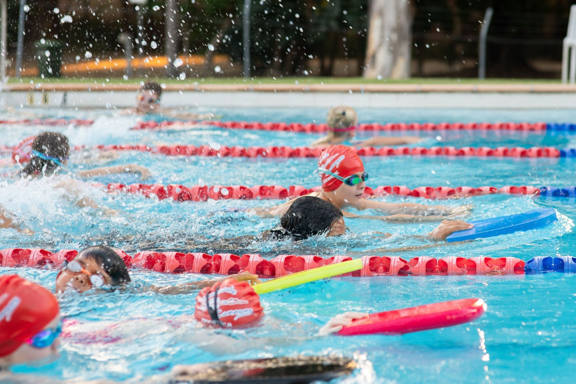 Swimmers in a pool, splashing water, using floats, red and blue lane markers.
