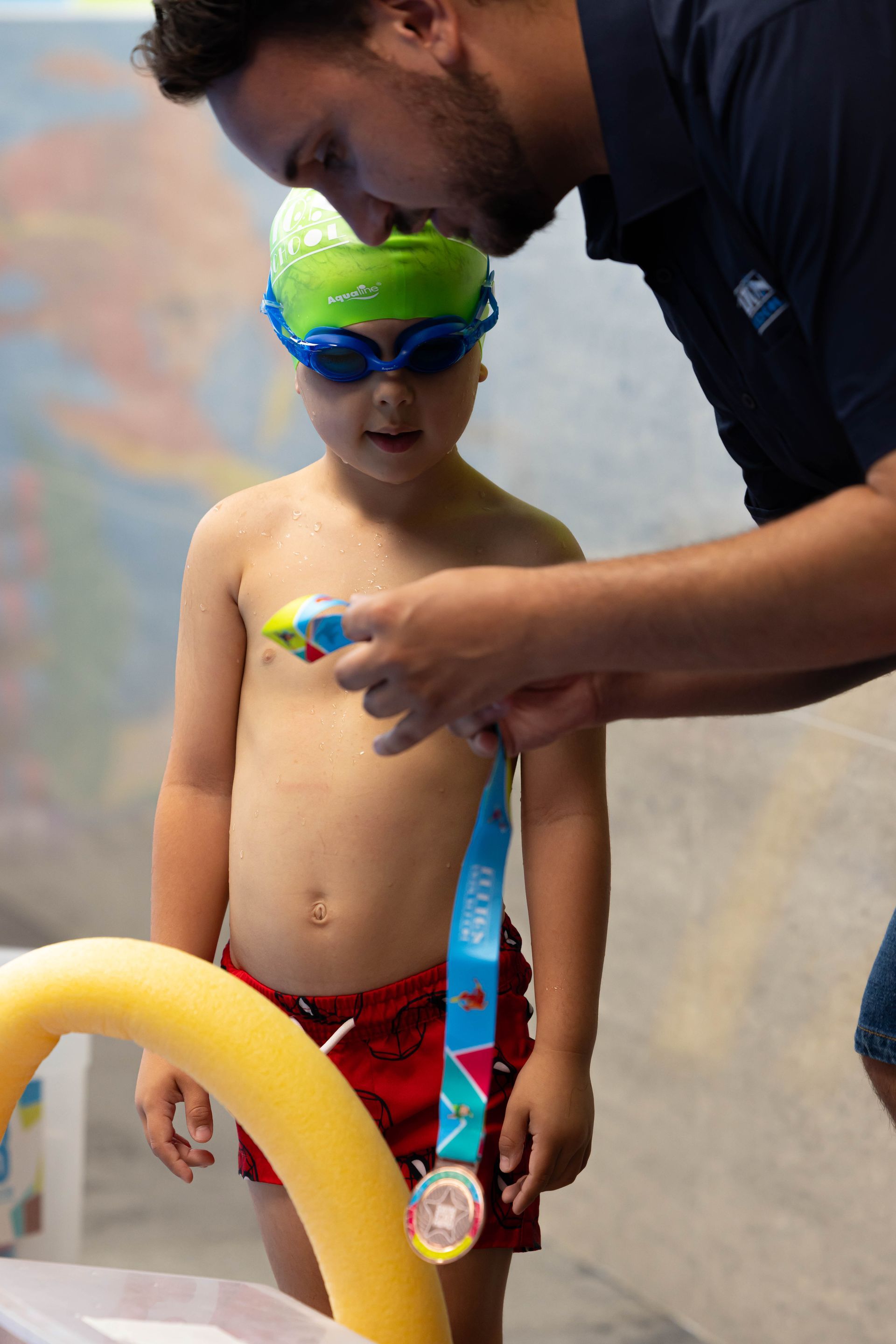 Child in swim goggles and red trunks by a pool, holding a medal