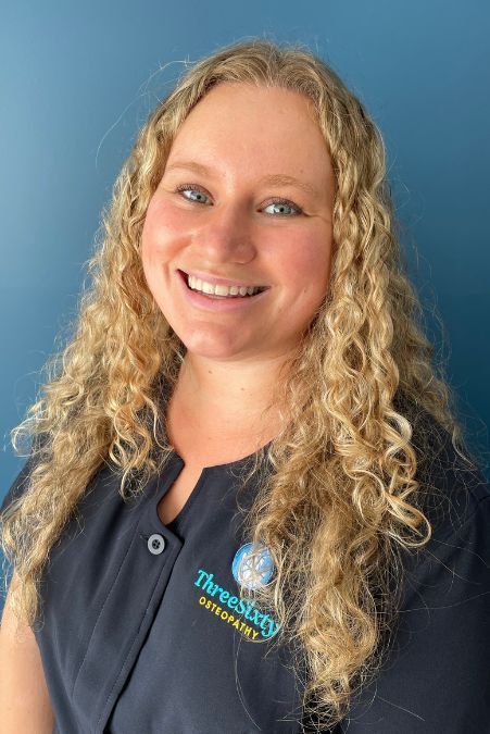 Woman with curly blonde hair smiles, wearing a dark blue uniform, in front of a blue wall.