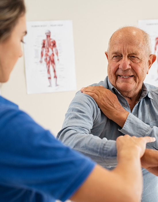 A healthcare worker examines an elderly man's shoulder, with a body chart on the wall.