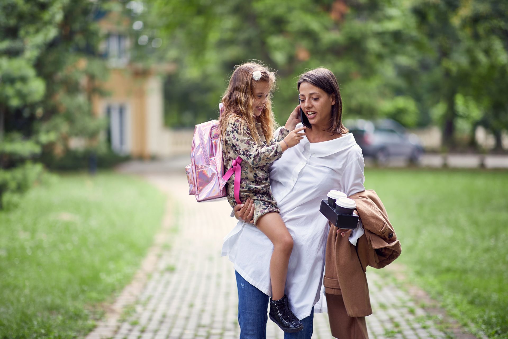 Woman carrying a girl on her shoulders, walking on path, school building, grass.