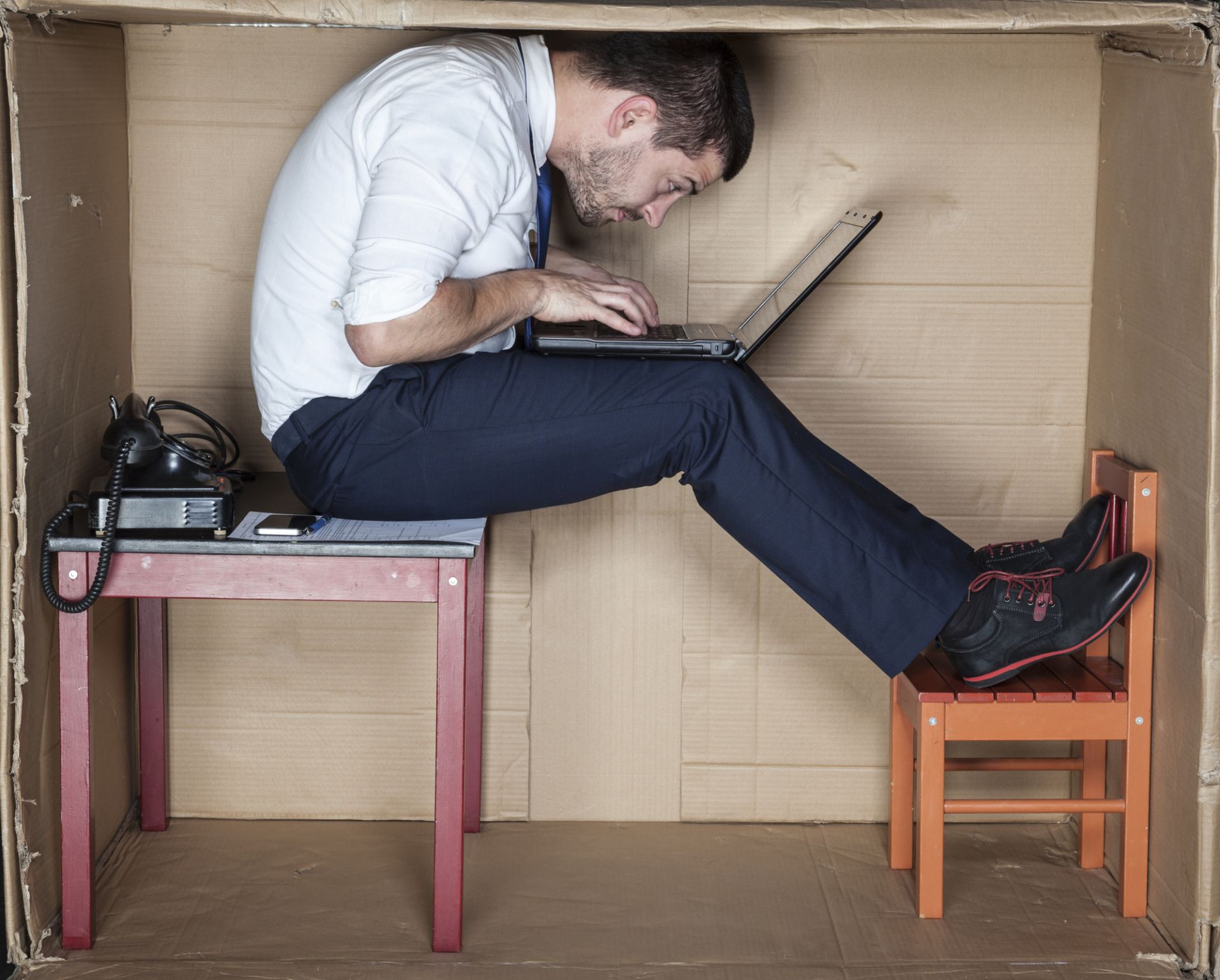 Man cramped in a cardboard box, hunched over laptop, sitting on a small table and chair, wearing business attire.