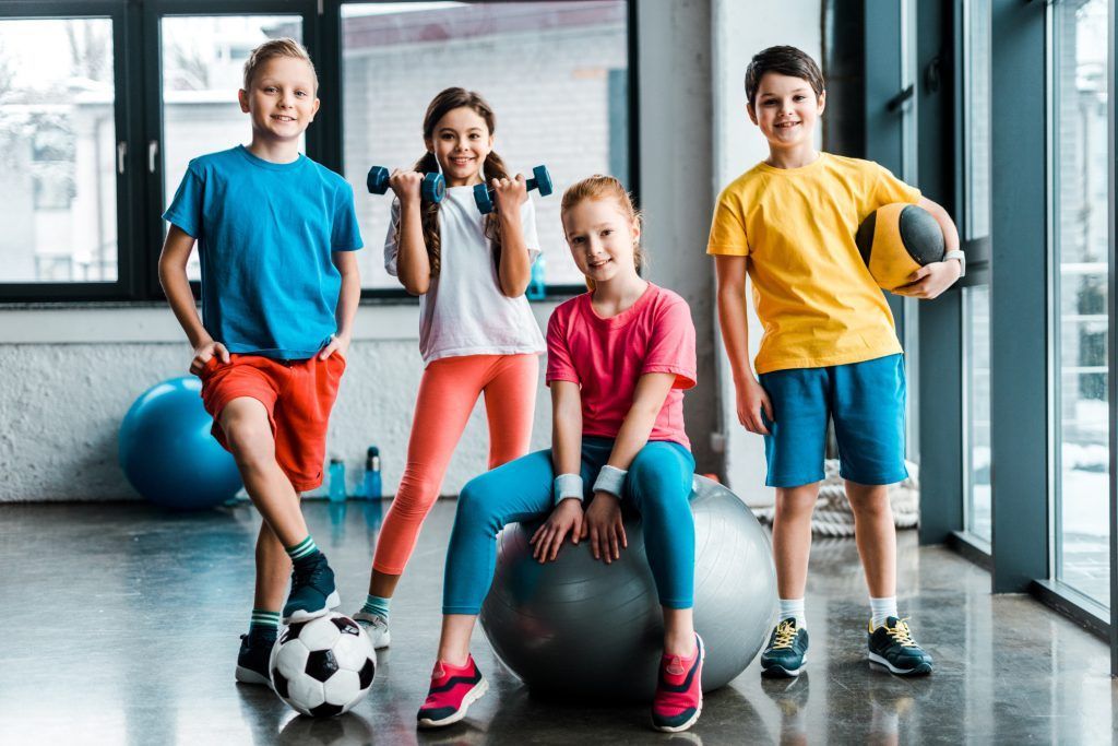 Four smiling kids in workout clothes, holding weights, ball, and posing in a gym.