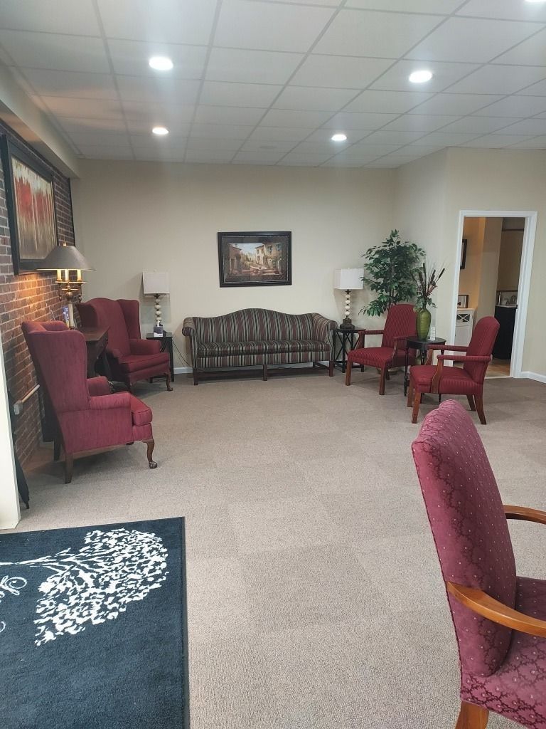 Waiting room with maroon chairs, a plaid sofa, framed art, and a potted plant on beige carpet.